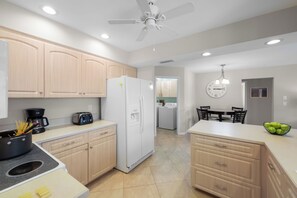 A modern kitchen with light wooden cabinets, a white refrigerator, a coffee maker, and a dining area with a clock.