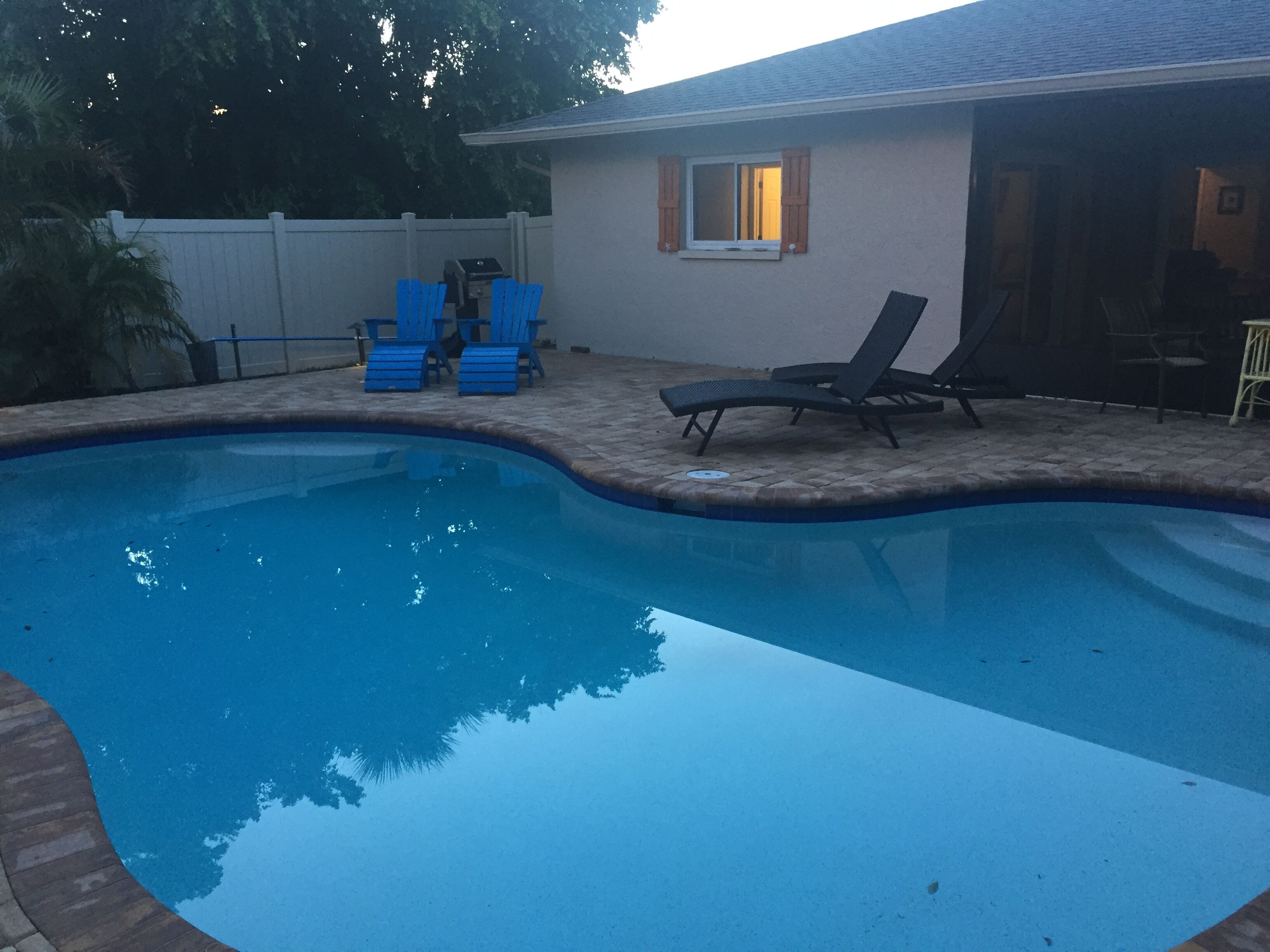 A swimming pool with a brick edge, a covered patio area with blue chairs, and a house with a window and door.