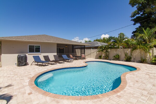 Une piscine à bordures courbes, entourée d’une terrasse carrelée. On y trouve des chaises longues et un barbecue.