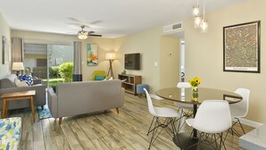 A modern living room with a sofa, a dining table with chairs, a ceiling fan, and a TV mounted on the wall.