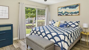 A bedroom with a bed, a ceiling fan, a window with curtains, a bedside table, and a wall-mounted painting.