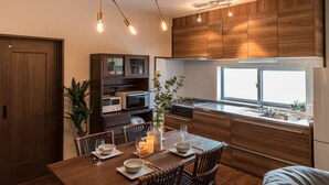 A modern kitchen with wooden cabinets, a dining table set for four, and a large window providing natural light. - BB Wakugawa Terrace (Nakijin)