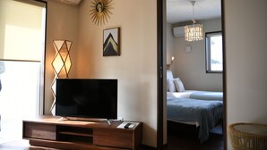 A hotel room with a TV on a wooden stand, a bed with a blue bedspread, a wall-mounted picture, and a sunburst decoration. - BB Wakugawa Terrace (Nakijin)