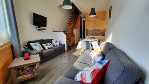 En moderne stue med en grå sofa, et fjernsyn monteret på væggen, og en trappe der f ører op til et køkken.