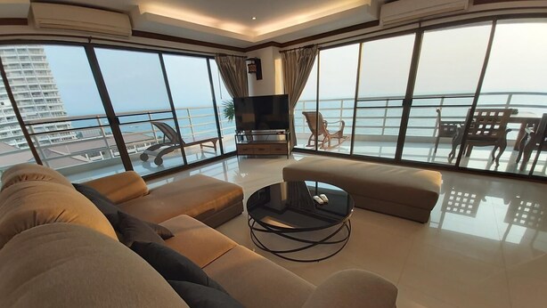 Un salon moderne avec un grand canapé, une table basse et un téléviseur. La pièce est dotée de grandes fenêtres qui offrent une vue sur la mer et un immeuble.