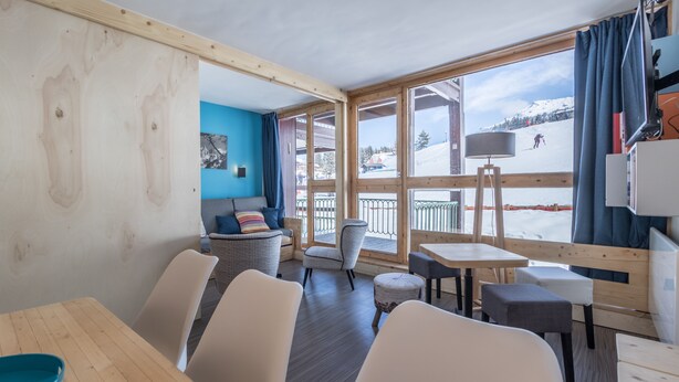Una habitación acogedora con techo de madera, ventanas amplias y vistas a una montaña nevada. Hay varias sillas y mesas, además de una pequeña mesa redonda con una bandeja azul.