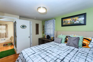 A bedroom with a bed, a door with a logo, a framed picture on the wall, and a bedside table with a lamp. - Game Day HQ w/ Fan Cave, Fire Pit + Stadium Views (Green Bay)