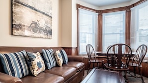 A living room with a brown leather sofa, patterned cushions, a wooden coffee table, and a dining table with chairs. There is a large painting on the wall and a window with blinds.