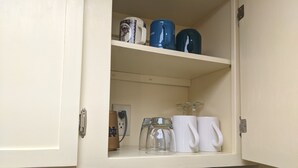A kitchen cabinet with a white interior, containing various kitchenware items like mugs, a glass pitcher, and a jar.
