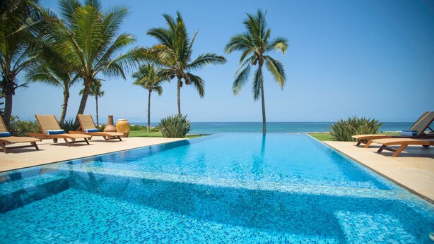 Une piscine à l’eau d’un bleu cristallin, entourée de chaises longues et de palmiers, avec vue sur l’océan.