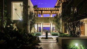 Un bâtiment moderne à deux étages, doté d’une entrée bien éclairée et entouré d’une végétation tropicale luxuriante.