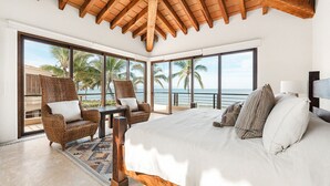 Une chambre spacieuse avec un grand lit, deux fauteuils en osier, une petite table et une vue sur l’océan grâce à de grandes fenêtres.
