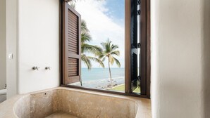 Une salle de bain dotée d’une grande baignoire, d’une fenêtre avec des volets en bois et offrant une vue sur la plage et les palmiers.