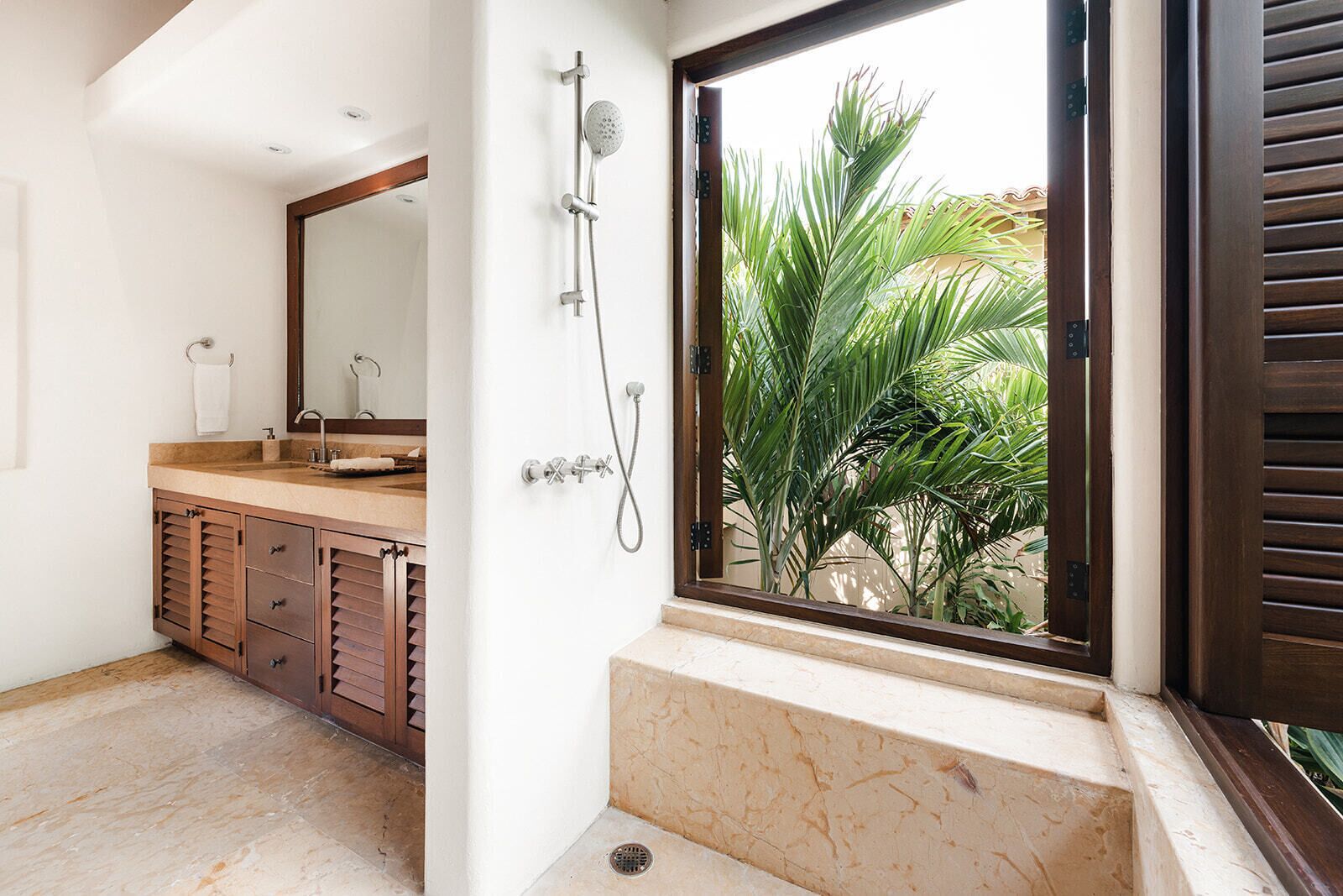 Un bagno con un mobile vanitario in legno, uno specchio grande, una doccia con soffione a mano e vista su palme attraverso una finestra.