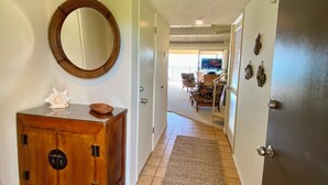 A hallway with a wooden cabinet, a mirror, and a door leading to a living area with a TV and furniture. - Stunning Beach Front Townhouse (Kihei)