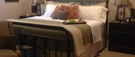 Un dormitorio ordenado con cama, dos mesitas de noche, una silla y una ventana que deja entrar luz natural.
