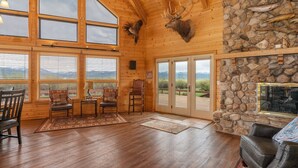 A log cabin interior with a stone fireplace, wooden walls, and large windows. - Teton Riverfront Retreat | Spacious, Peaceful, Perfect for Family Adventures (Victor)