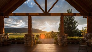 A wooden deck with stone pillars, a view of a sunset over a landscape, and a table with chairs. - Teton Riverfront Retreat | Spacious, Peaceful, Perfect for Family Adventures (Victor)