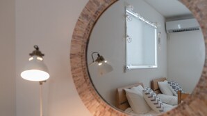 Een ronde spiegel met houten lijst die een bed weerspiegelt met witte en gemêleerde kussens, een bedlamp en een raam met zonwering.