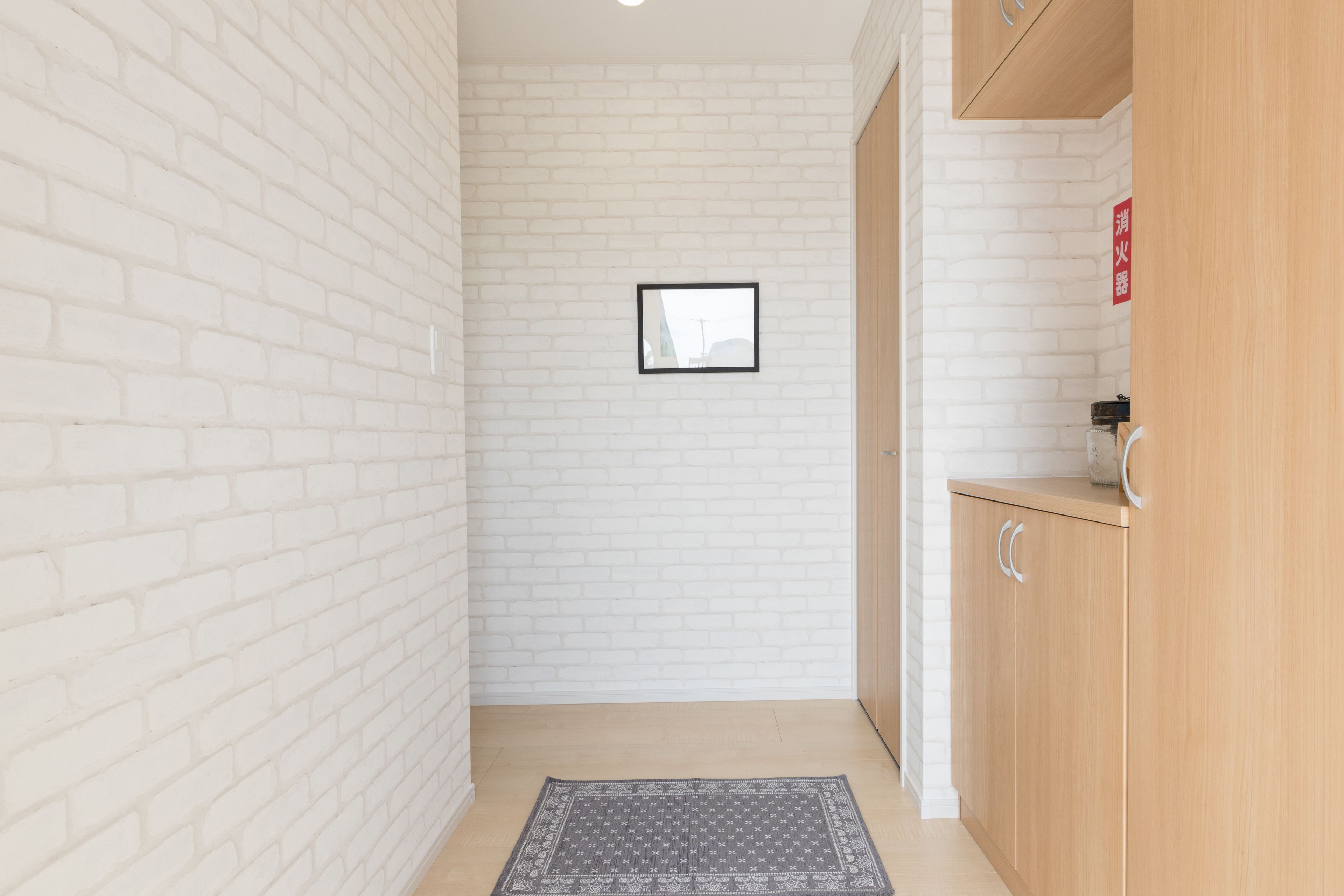 A narrow hallway with white brick walls, a framed picture on the wall, a wooden cabinet with a red sign, and a patterned rug on the floor.