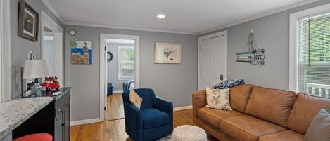 A living room with a brown leather sofa, a blue armchair, and a coffee table. There is a kitchen area with a counter and stools. The room has a gray wall and a wooden floor.