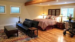Een slaapkamer met een groot bed, een houten plafond, twee bedlampjes, een klein tafeltje, een bankje en een vloerkleed met een patroon.