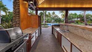 Een buitenkeuken met een barbecue, een spoelbak en uitzicht op een zwembad en een tropisch landschap.