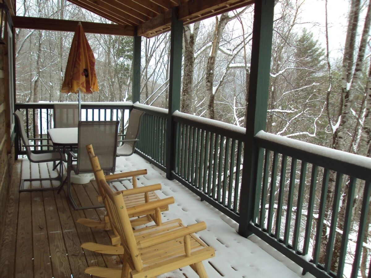A wooden deck with a table and chairs, a hanging umbrella, and a view of snow-covered trees.