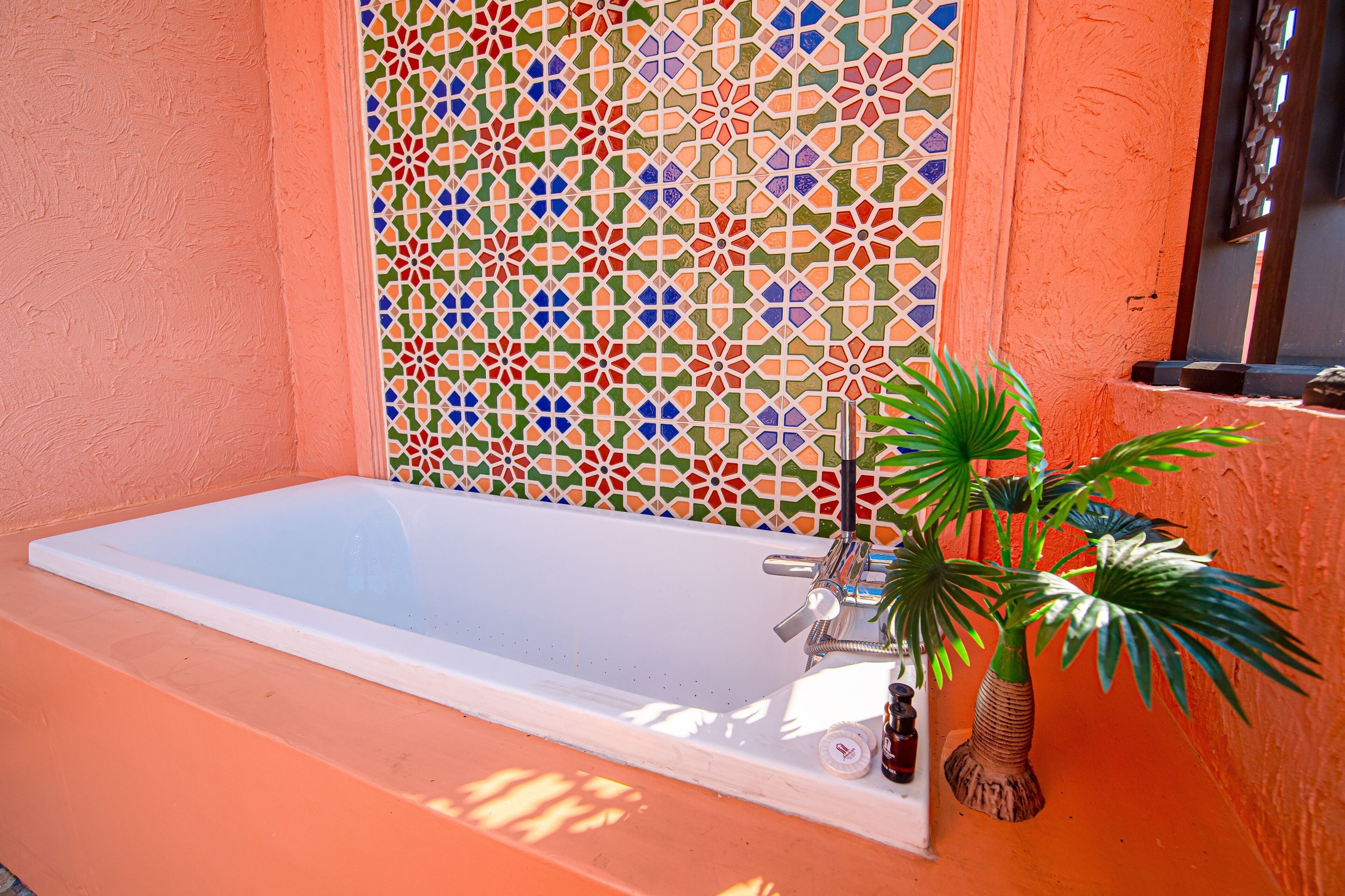 A bathtub with a colorful mosaic wall behind it, a potted plant beside the tub, and a window on the right.