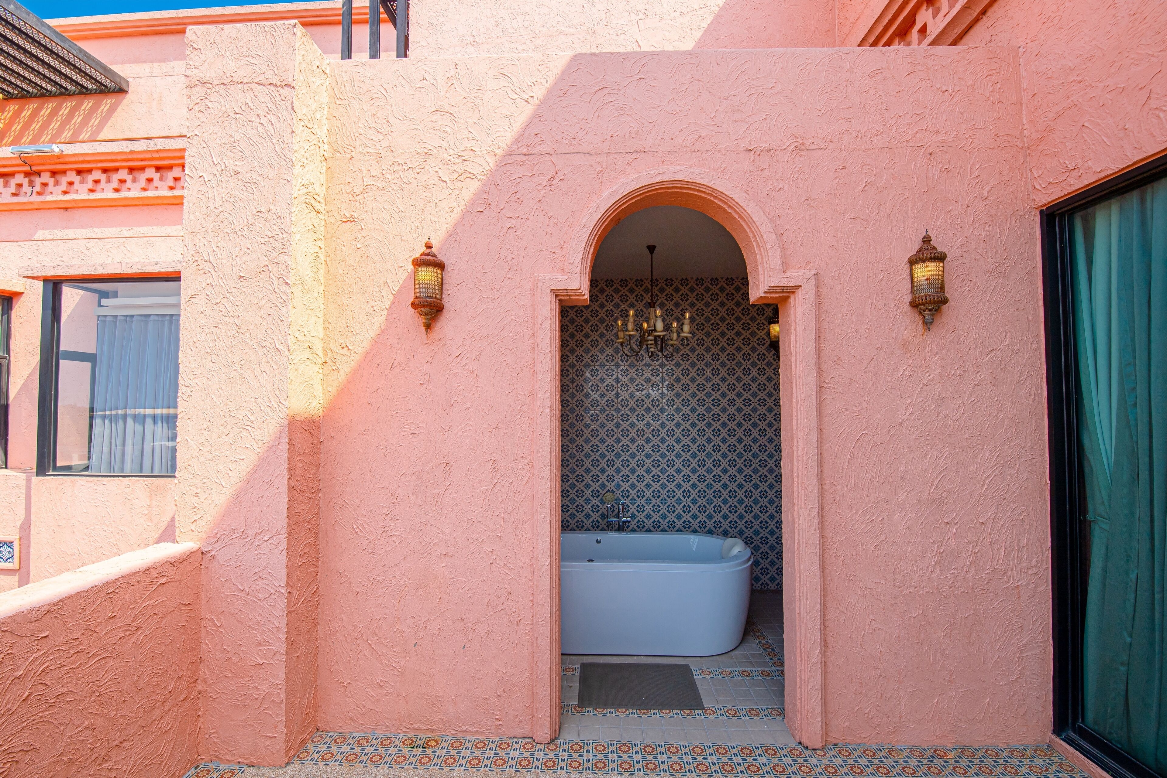 Un baño con paredes rosas, una bañera blanca, baldosas estampadas y dos faroles colgantes.
