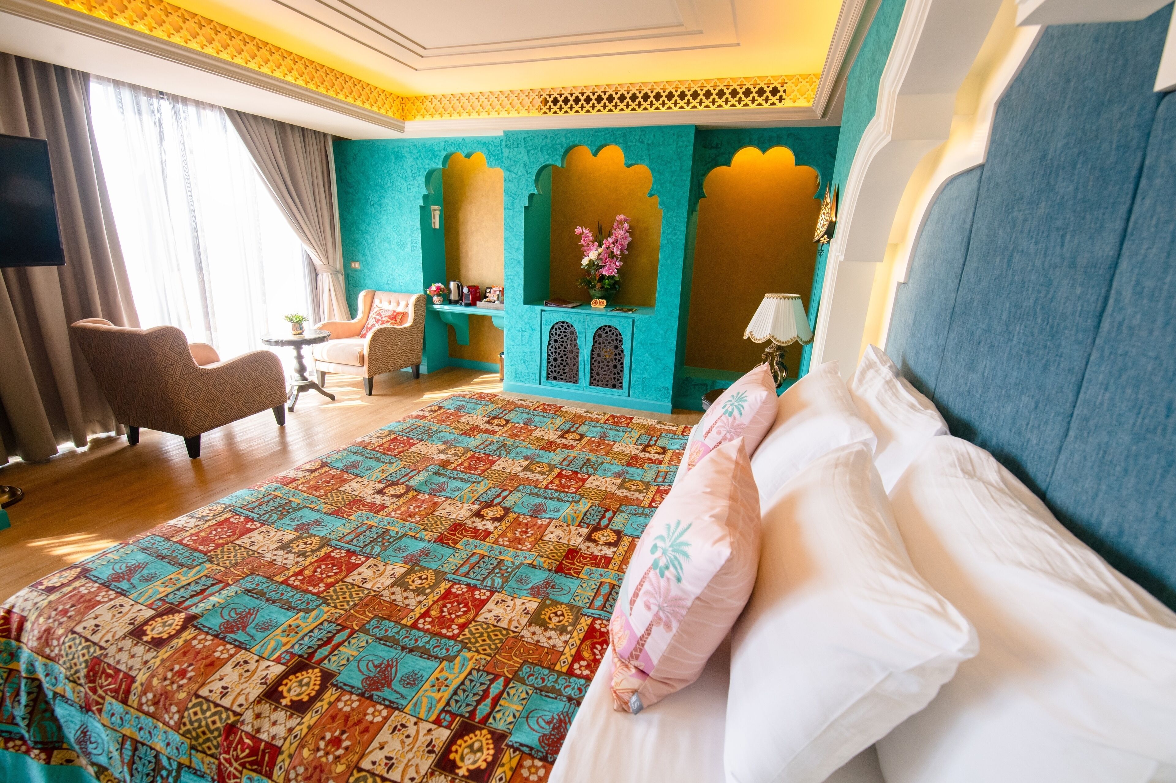 A hotel room with a large bed, a sitting area with a chair and a small table, and a decorative alcove with a vase of flowers.