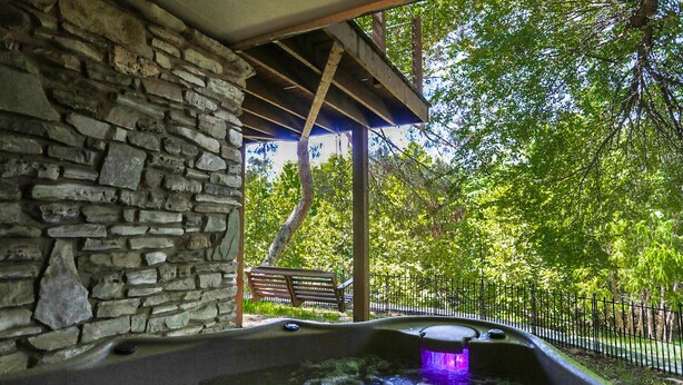 Eine Steinsäule, eine Holzpergola und ein Wasserspiel mit einer Bank und einem Zaun in einer Parkanlage.