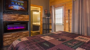 A cozy room with a large TV mounted on a wooden wall, a fireplace, a bed with a quilt, and a door leading to another room. - 390 Reviews ❤️ Mtn Top Getaway! Views, 2 Decks, Hot tub, Recliners, Privacy (Pigeon Forge)