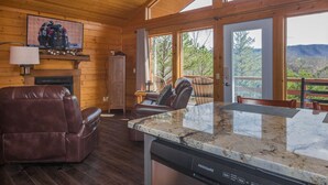A wooden cabin interior with a fireplace, leather recliner, granite countertop, and large windows overlooking a mountainous landscape. - 390 Reviews ❤️ Mtn Top Getaway! Views, 2 Decks, Hot tub, Recliners, Privacy (Pigeon Forge)