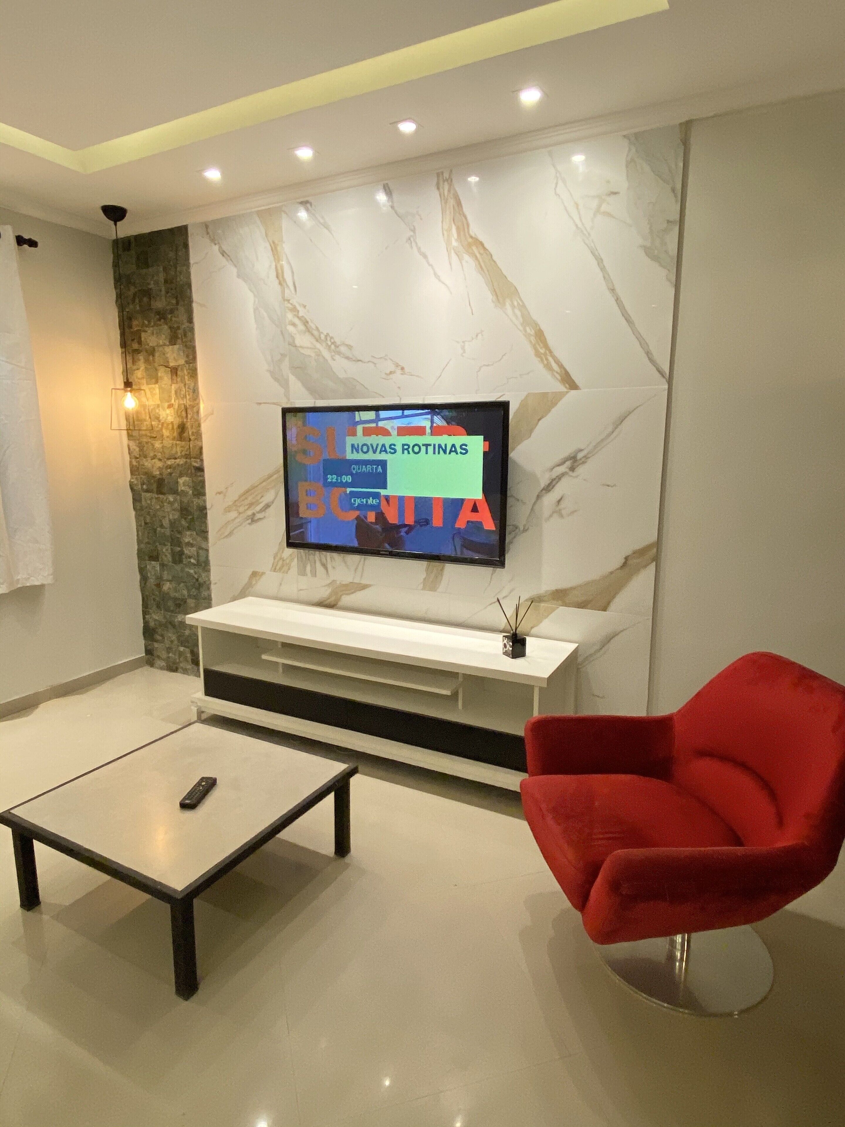 A modern living room with a flat-screen TV mounted on a marble wall, a red chair, a coffee table, and a remote control on the table.