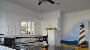 Une cuisine avec des meubles blancs, un plan de travail noir et un tableau représentant un phare.