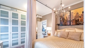 A bedroom with a large bed, a cityscape painting, a chandelier, and a walk-in closet.