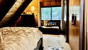 A cozy attic bedroom with a bed, a nightstand, a lamp, a window with a view of trees, and a fire extinguisher. - Perfect Sierra Cabin Retreat - Near Rivers, Lakes, and Mountains (Twin Bridges)
