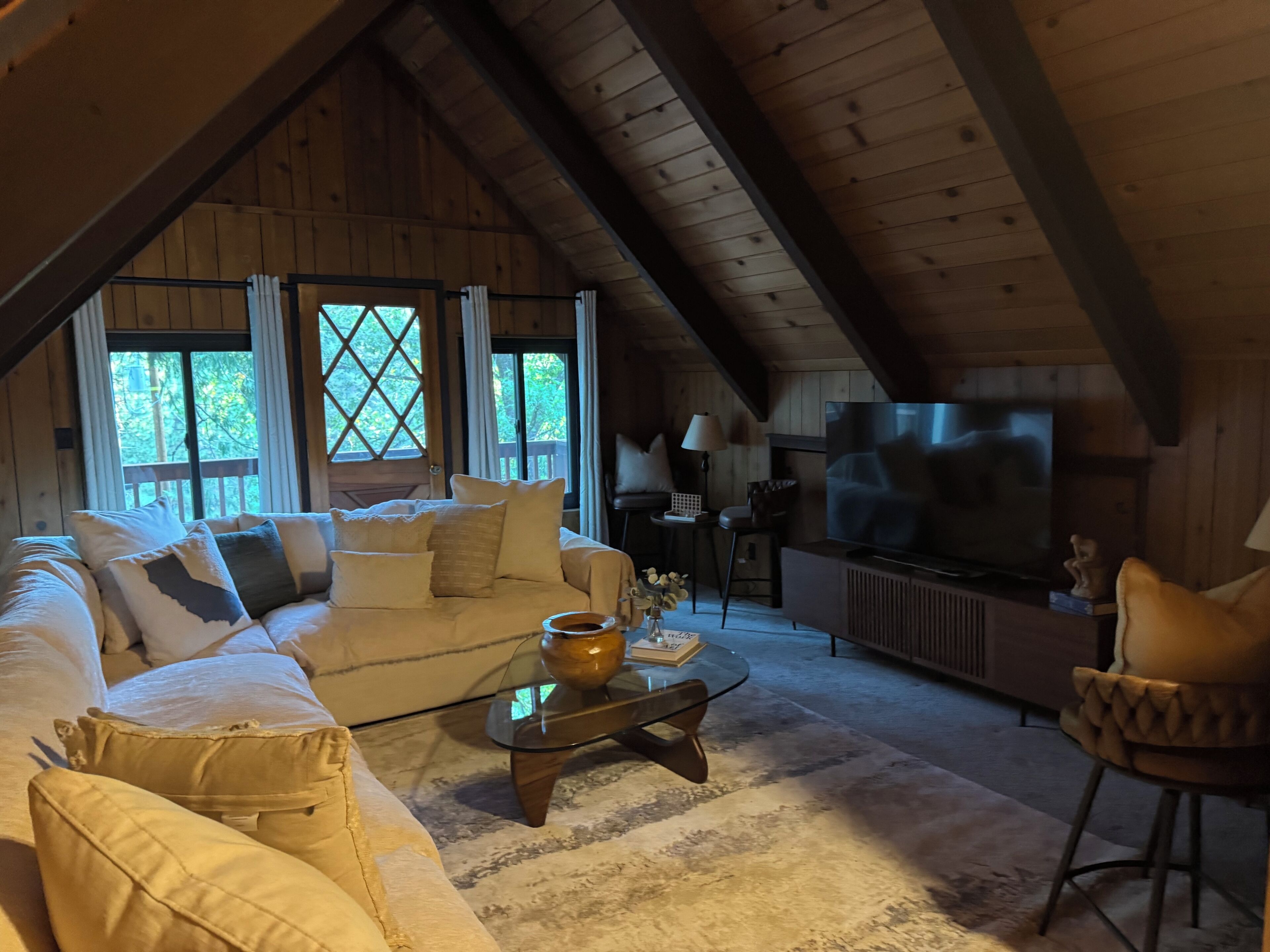 An attic room with a wooden ceiling, a sofa, a glass coffee table, a television, and a view of trees through large windows.