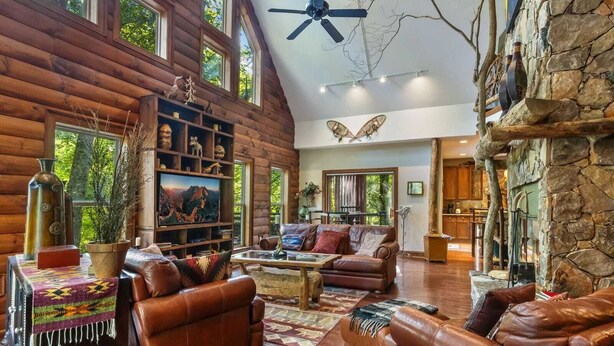 A spacious log cabin interior with a fireplace, leather furniture, a stone wall, and a ceiling fan.