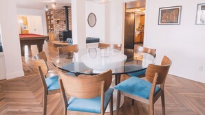 Een eetgedeelte met een tafel met glazen blad, houten stoelen met blauwe kussens en een open haard van baksteen op de achtergrond.