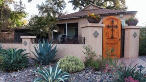 A house with a wooden front door, a balcony with a railing, and a garden with various plants and rocks.