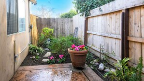 A small, enclosed garden area with a wooden fence, a potted plant with pink flowers, and various shrubs and rocks.