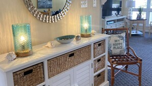 Een woonkamer met een witte console tafel, rieten manden, een ronde spiegel, een televisie en een stoel met kussen.