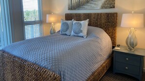 Een net opgemaakt bed met een gebreide deken, twee bijpassende kussens en een hoofdbord met een geweven patroon. Aan weerszijden van het bed staan twee nachtlampjes, en erboven hangt een schilderij van een strandgezicht.