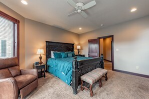 A bedroom with a large bed, a wooden bench, a brown leather chair, a ceiling fan, and a door leading to another room.