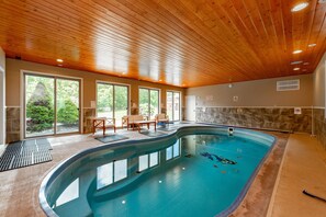 An indoor swimming pool with a wooden ceiling, large windows, and a seating area with chairs and a table.
