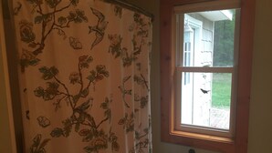 Un baño con cortina de ducha estampada con motivos florales, una ventana que ofrece vista a una casa y un pájaro, y un dispensador de papel higiénico en la pared.