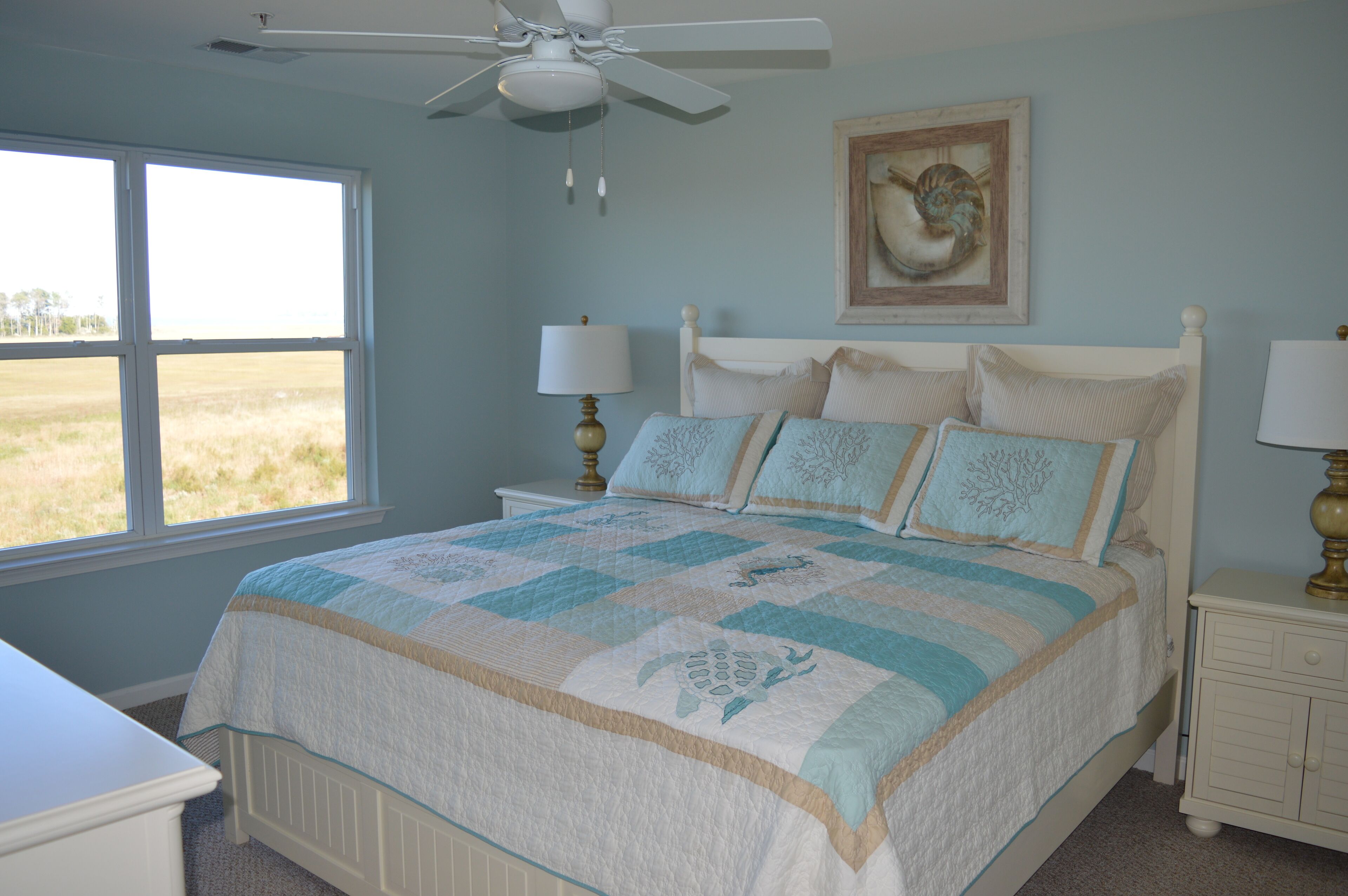 A bedroom with a large window, a ceiling fan, a bed with a quilt, bedside tables with lamps, and a framed picture on the wall.