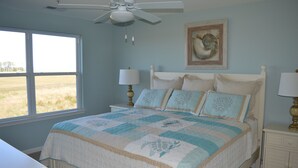 A bedroom with a large window, a ceiling fan, a bed with a quilt, bedside tables with lamps, and a framed picture on the wall. - 2 story condo w/ Stunning Views and Many Amenities (Ocean View)